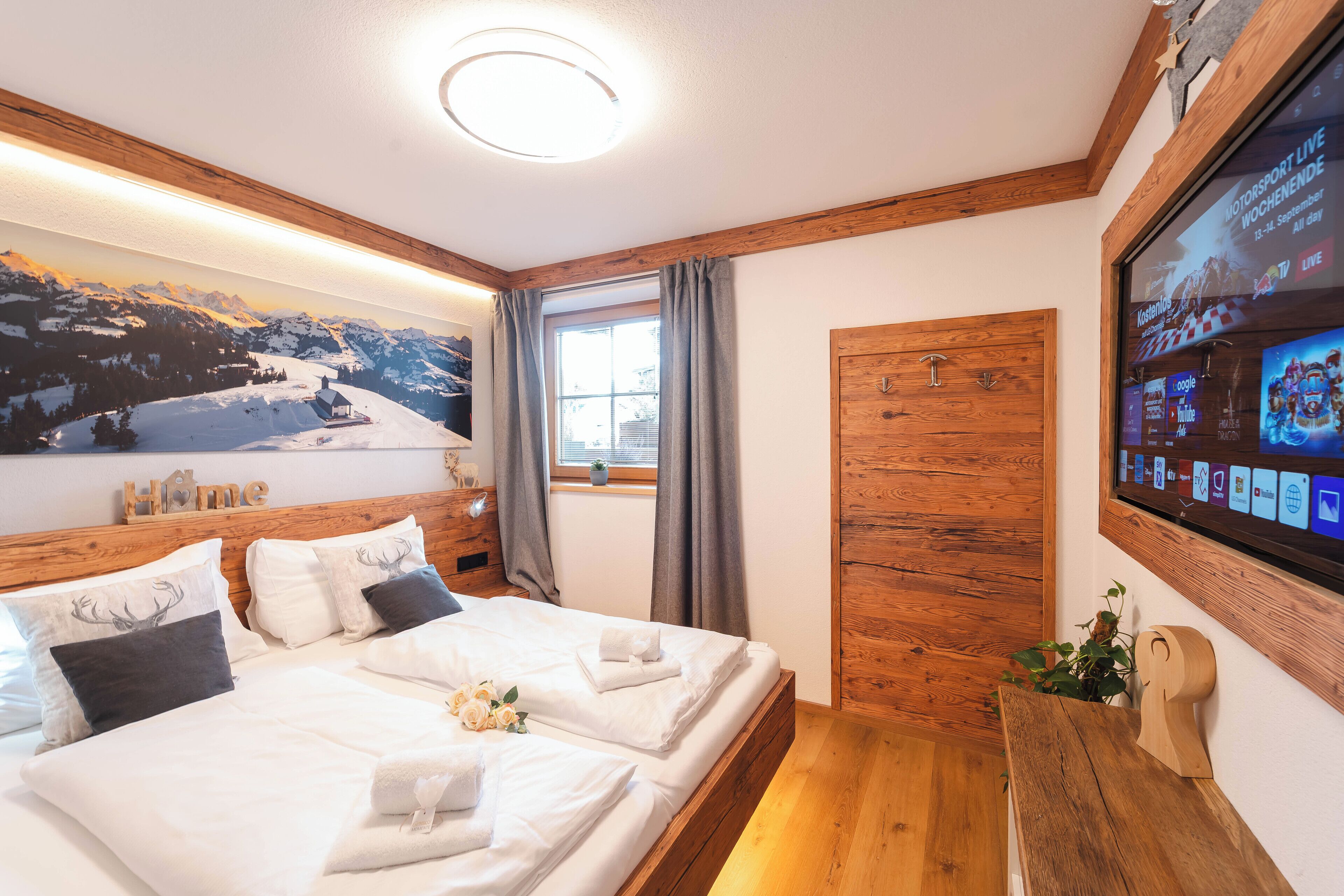 Habitación de hotel con cama, cabecero de madera, un ventanal con cortinas, televisor montado en la pared y un cartel con vistas a la montaña.