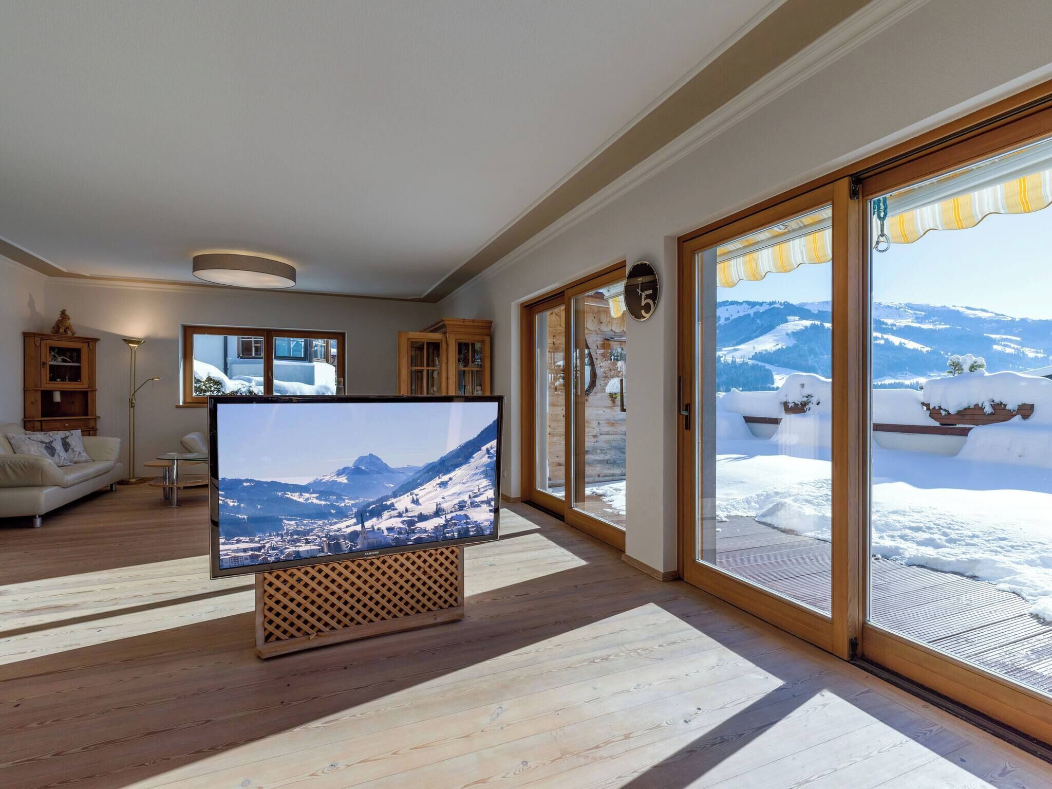Una sala de estar moderna con un televisor de pantalla plana grande, piso de madera y vistas a un paisaje montañoso nevado a través de grandes puertas correderas de vidrio.
