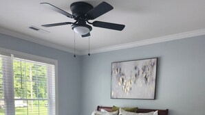Ein Schlafzimmer mit einem Bett, einem Deckenventilator, einem Fenster mit Jalousien, einem Teppich und einem Nachttisch mit Lampe.