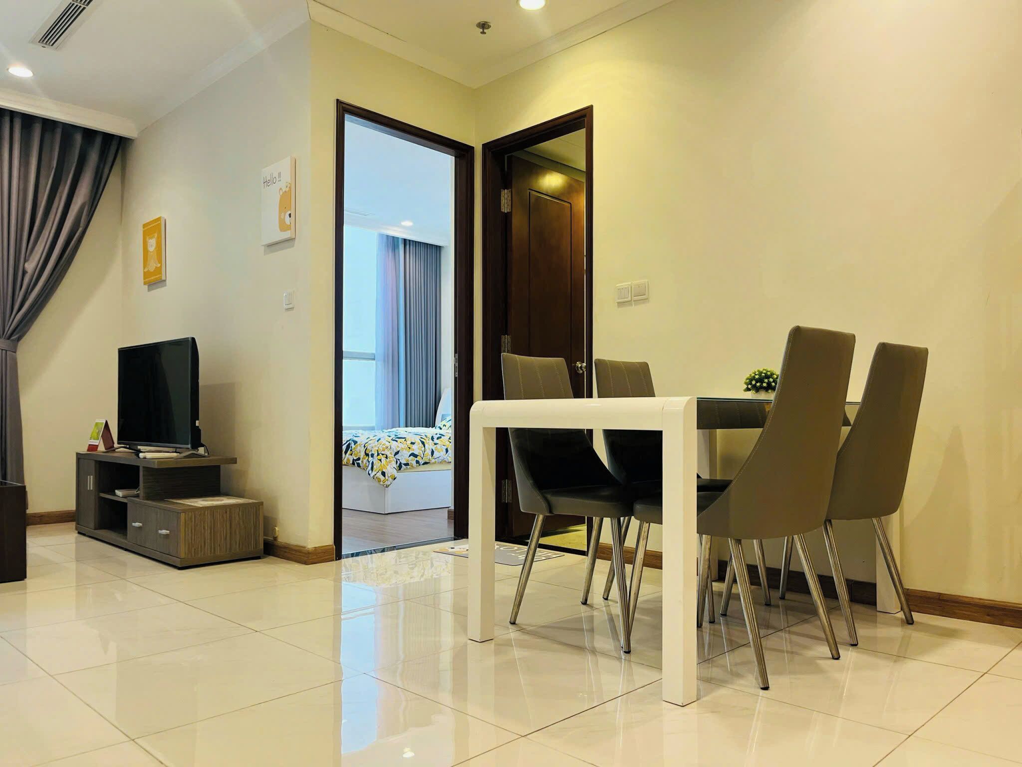 A modern living room with a flat-screen TV, a dining table with four chairs, and a doorway leading to a bedroom.