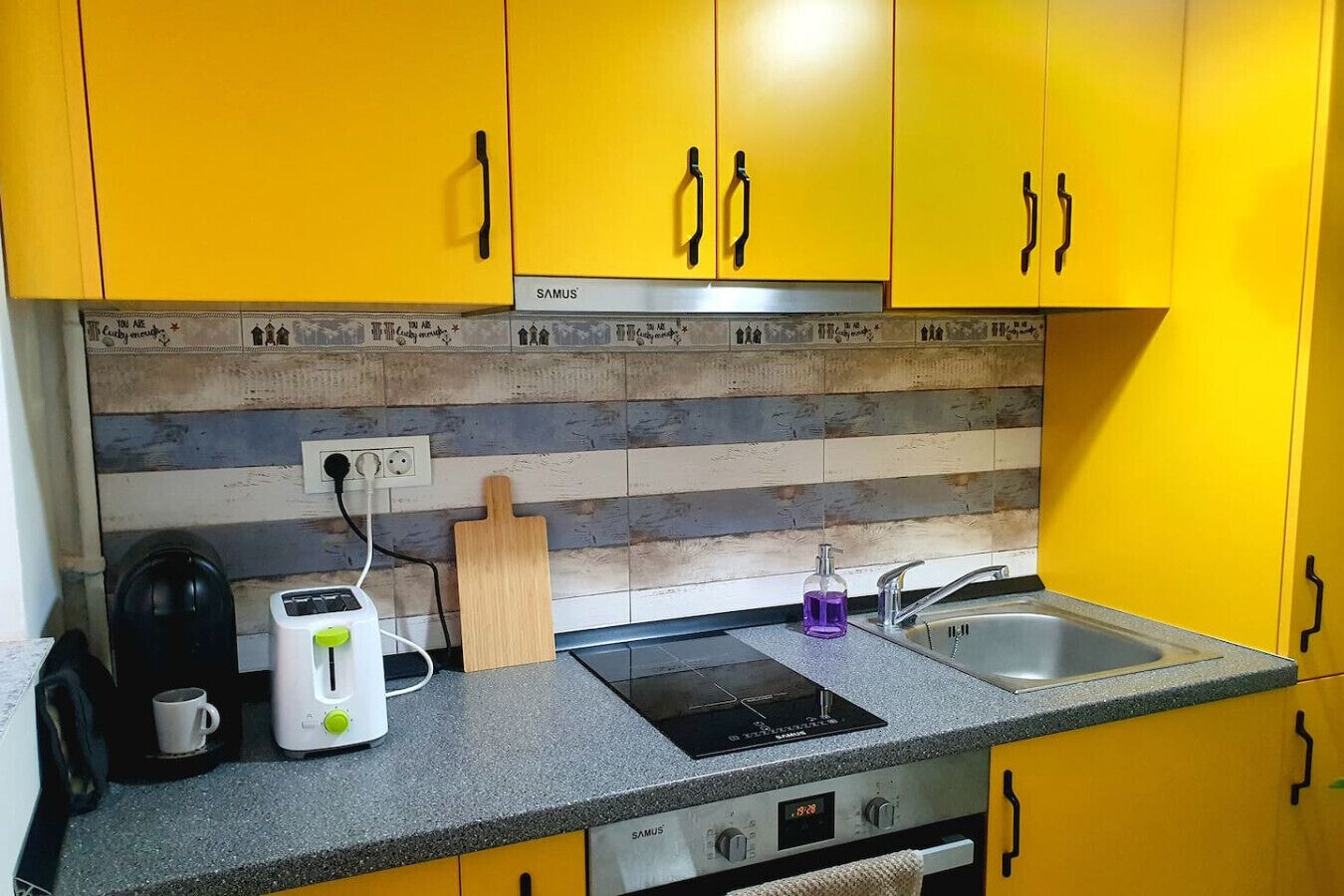 Une cuisine avec des meubles de couleur jaune, un pare-feu en bois, un grille-pain, une bouilloire, une planche à découper et un évier.