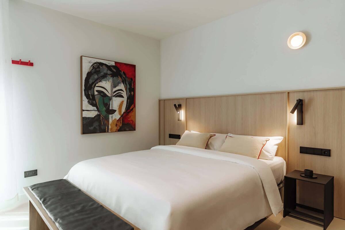 Una habitación de hotel moderna con una cama grande, un banco al pie de la cama, un cuadro en la pared y una mesita de noche con una lámpara.