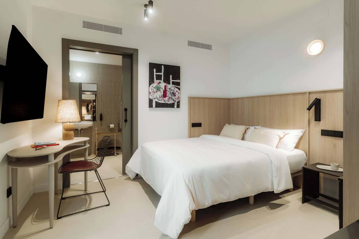 Una habitación de hotel moderna con cama, mesita de noche, silla, escritorio y un cuadro en la pared.