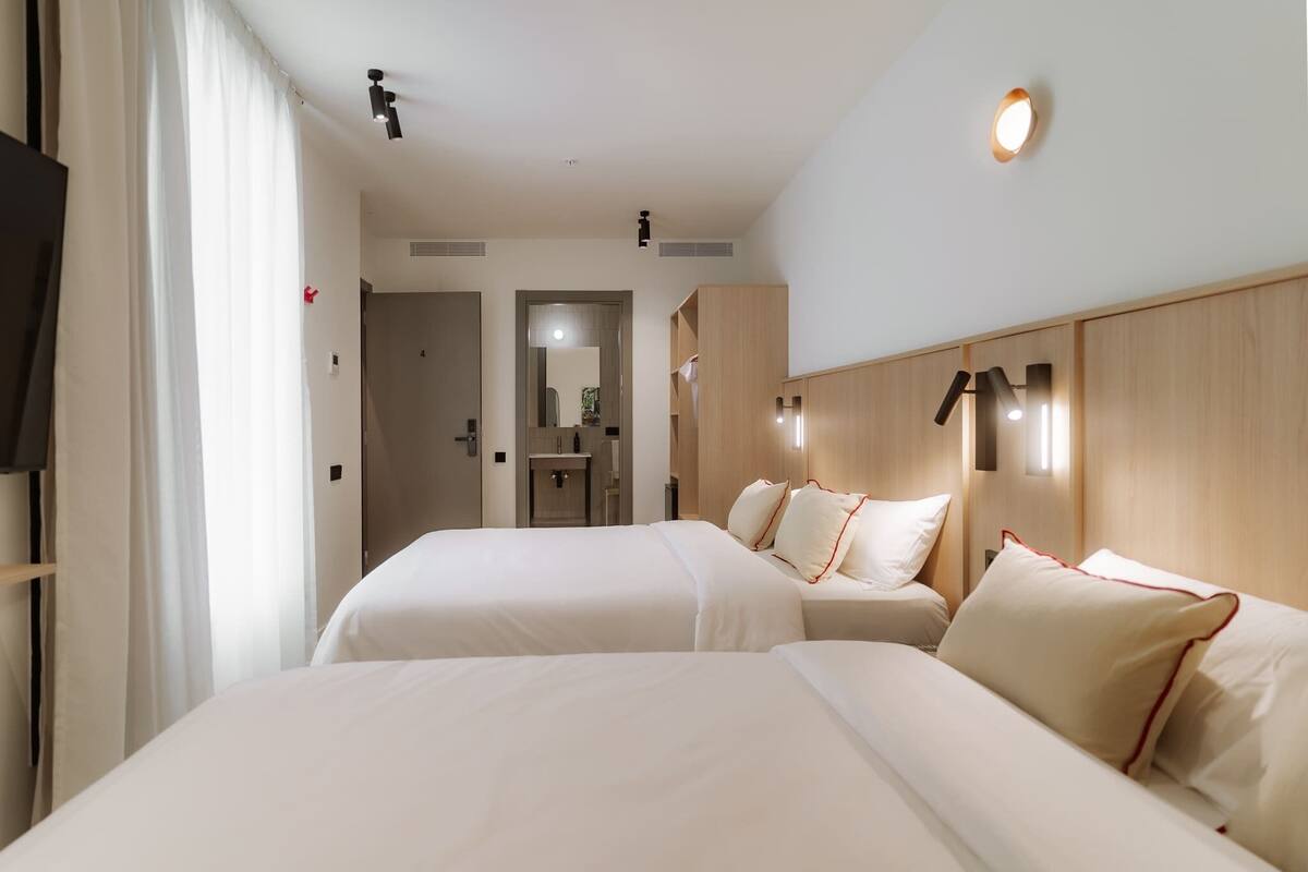 Habitación de hotel con dos camas, cabecera de madera, un espejo y un baño pequeño visible a través de una puerta abierta.