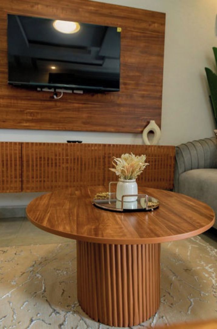A modern living room with a wooden TV wall mount, a round wooden coffee table with a vase of dried flowers, and a grey sofa.