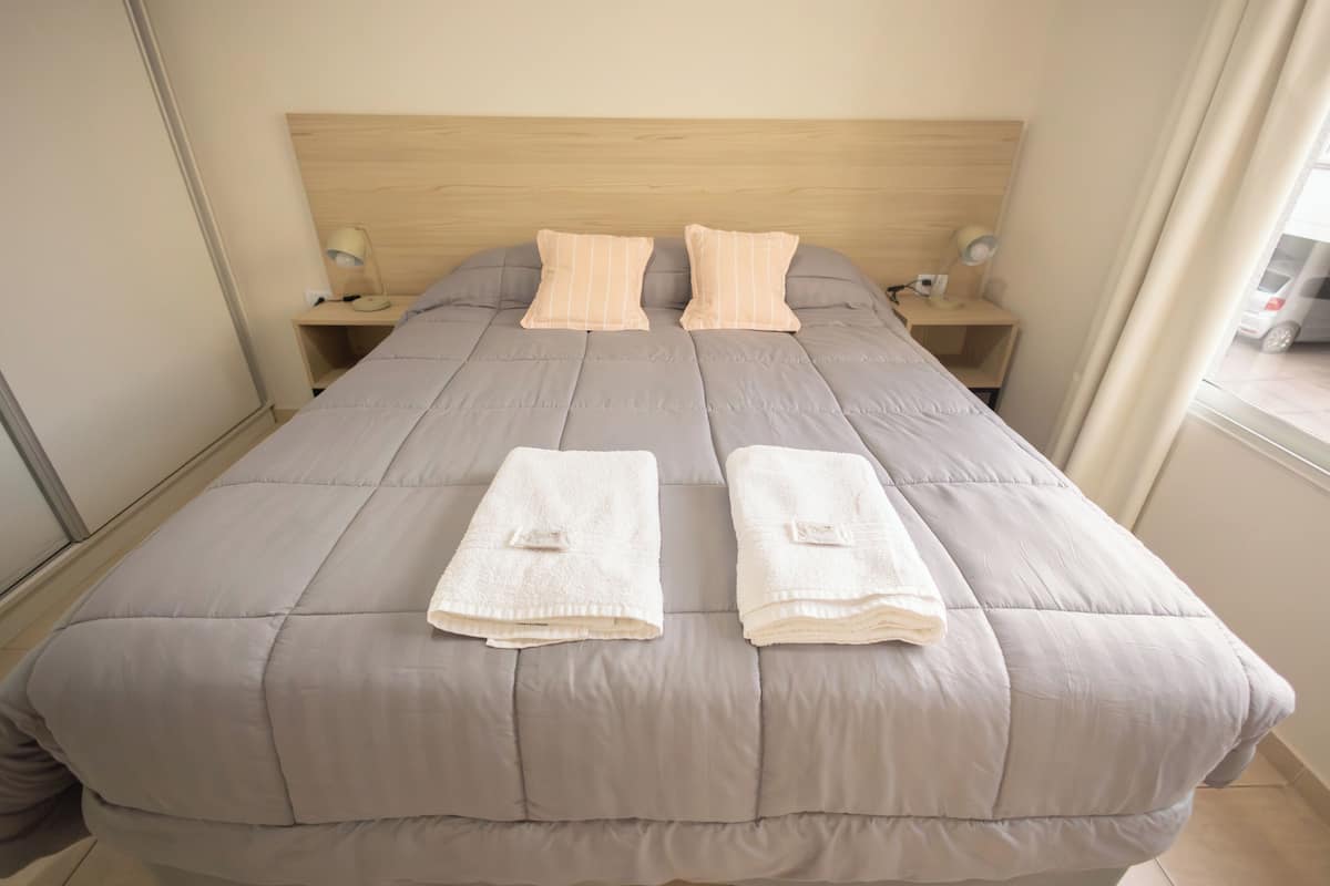 A neatly made bed with a grey duvet, two pillows, and two folded towels. There are bedside tables with lamps and a window with curtains.