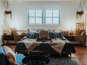 Quarto com cama grande, janela com persianas, mesa de cabeceira e planta pendente.