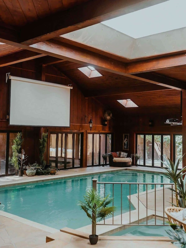 Piscina interior com teto de madeira e claraboias, rodeada por plantas em vasos e uma área de estar com vista para o exterior.