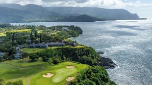 A golf course with bunkers and a clubhouse, surrounded by lush greenery and overlooking a large body of water with mountains in the background.