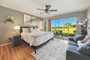 Un dormitorio con cama, mesita de noche, lámpara, ventilador de techo, obra de arte y vista a un campo de golf a través de amplias ventanas.