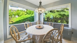 A dining area with a round table, six chairs, and a chandelier. It features large windows with a view of lush greenery.