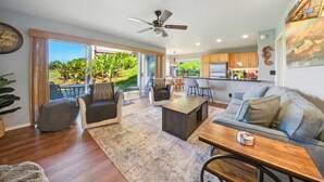 A spacious living area with a large window, a ceiling fan, and a kitchen in the background.