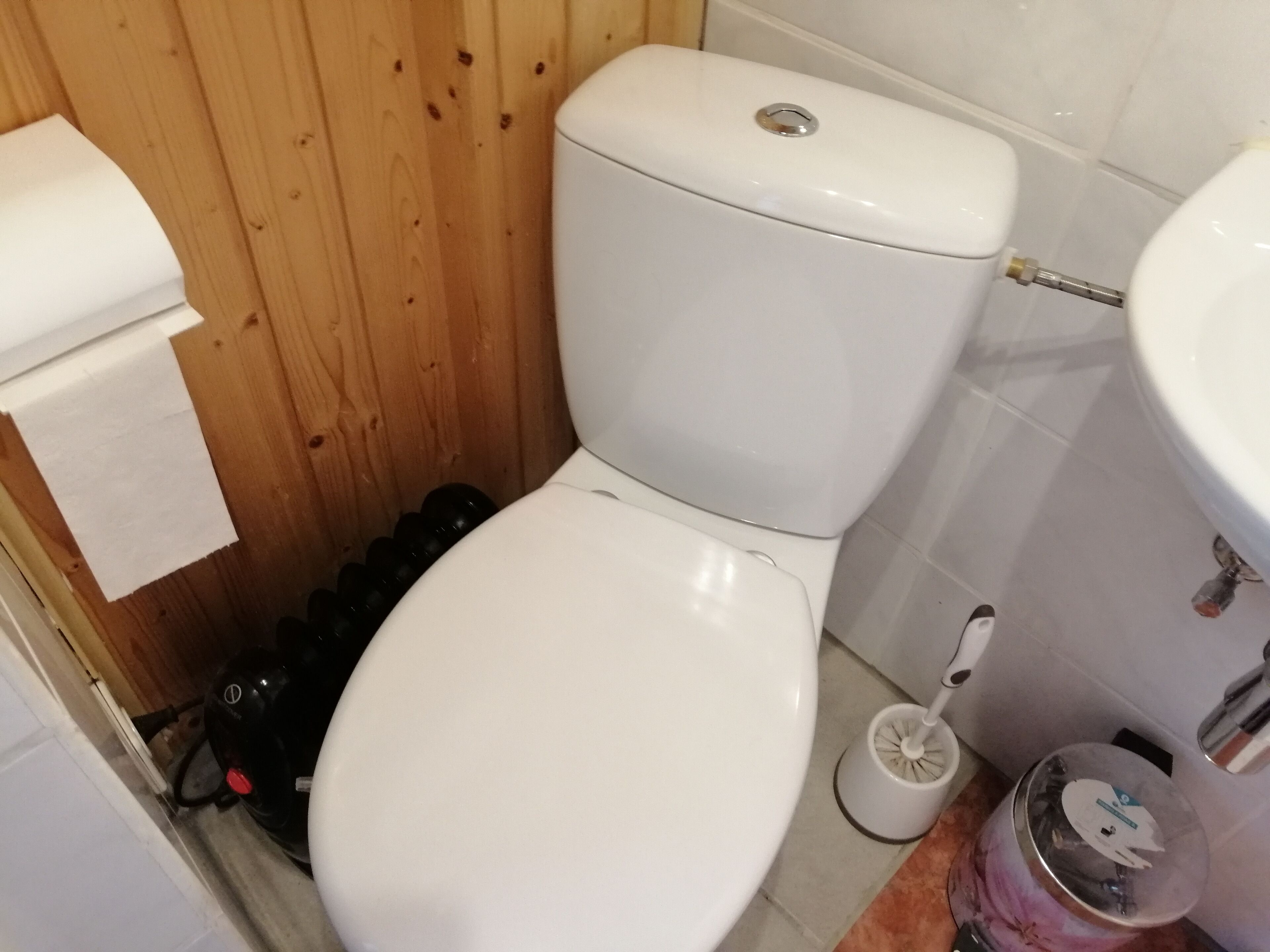 Pieni kylpyhuone, jossa on valkoinen wc-istuin, puinen seinä, valkoinen pesuallas ja wc-harja.
