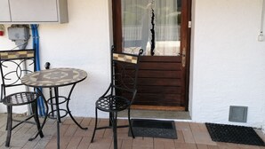 A small outdoor seating area with a table and chairs, a wooden door with a glass panel, and a white wall with a power outlet. - Le Logis du bChalet Perce-Neige Route de la Cierne 91Les Paccots 1619 (Châtel-Saint-Denis)