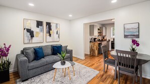 Ein modernes Wohnzimmer mit grauem Sofa, einem kleinen runden Tisch und einem Essbereich mit Holztisch und Stühlen.