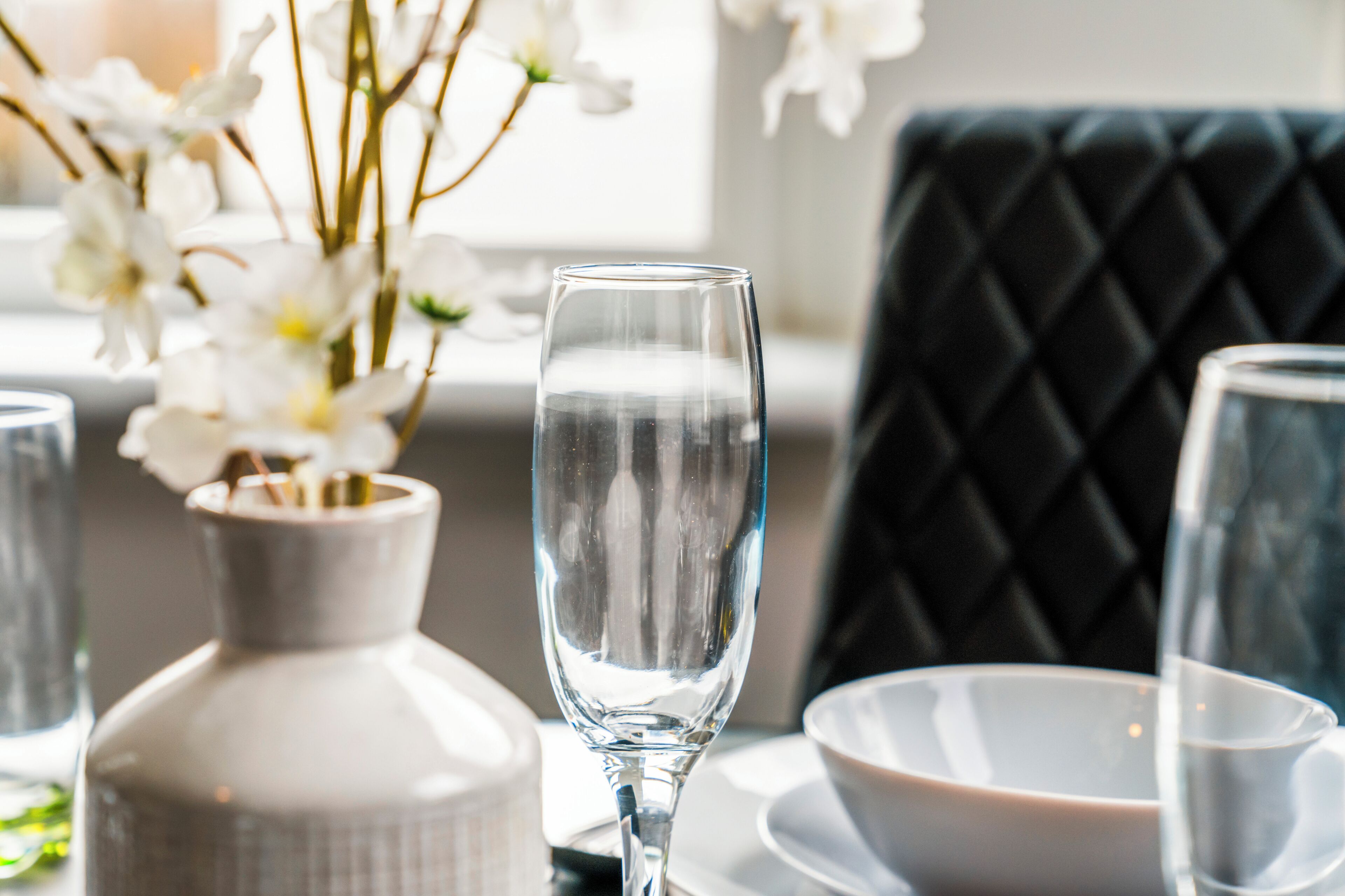 Une table à manger avec un plateau en verre transparent, un vase blanc orné de fleurs et une chaise noire avec un coussin.