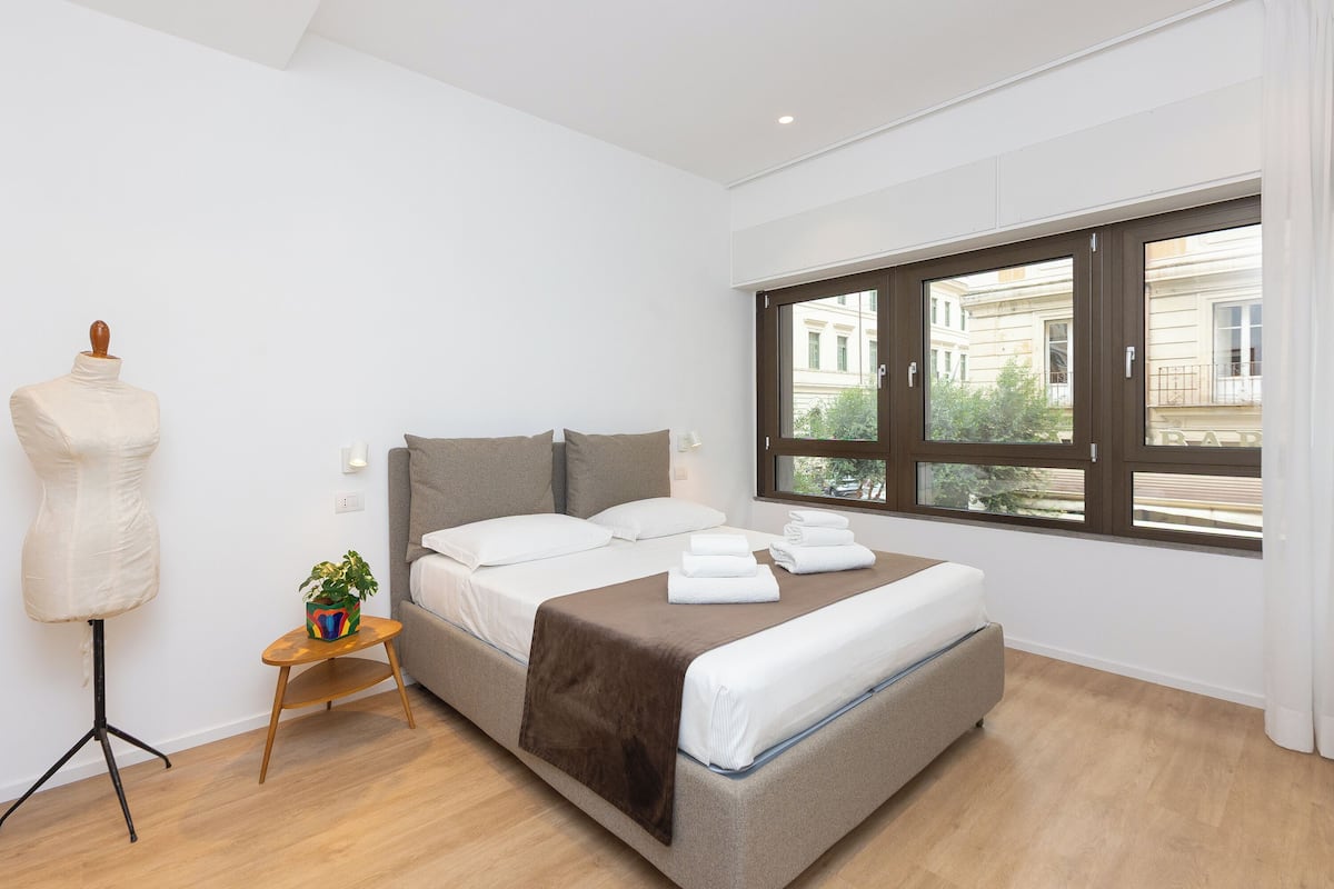 Ein modernes Schlafzimmer mit einem großen Bett, einem kleinen Nachttisch mit Pflanze und einem Fenster mit Blick auf Gebäude.