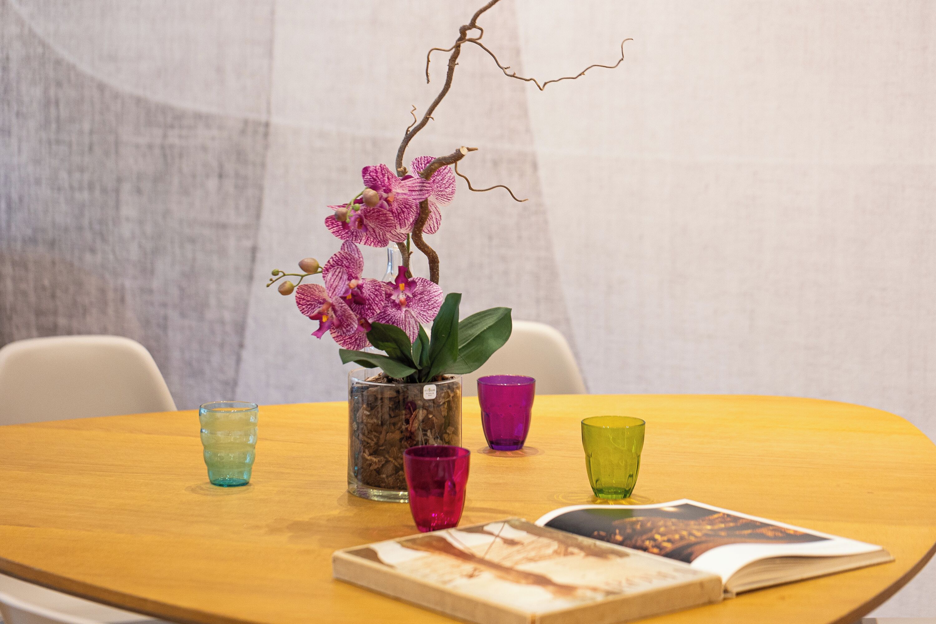 Una mesa redonda de madera con una orquídea en maceta, un libro y tres vasos de colores.