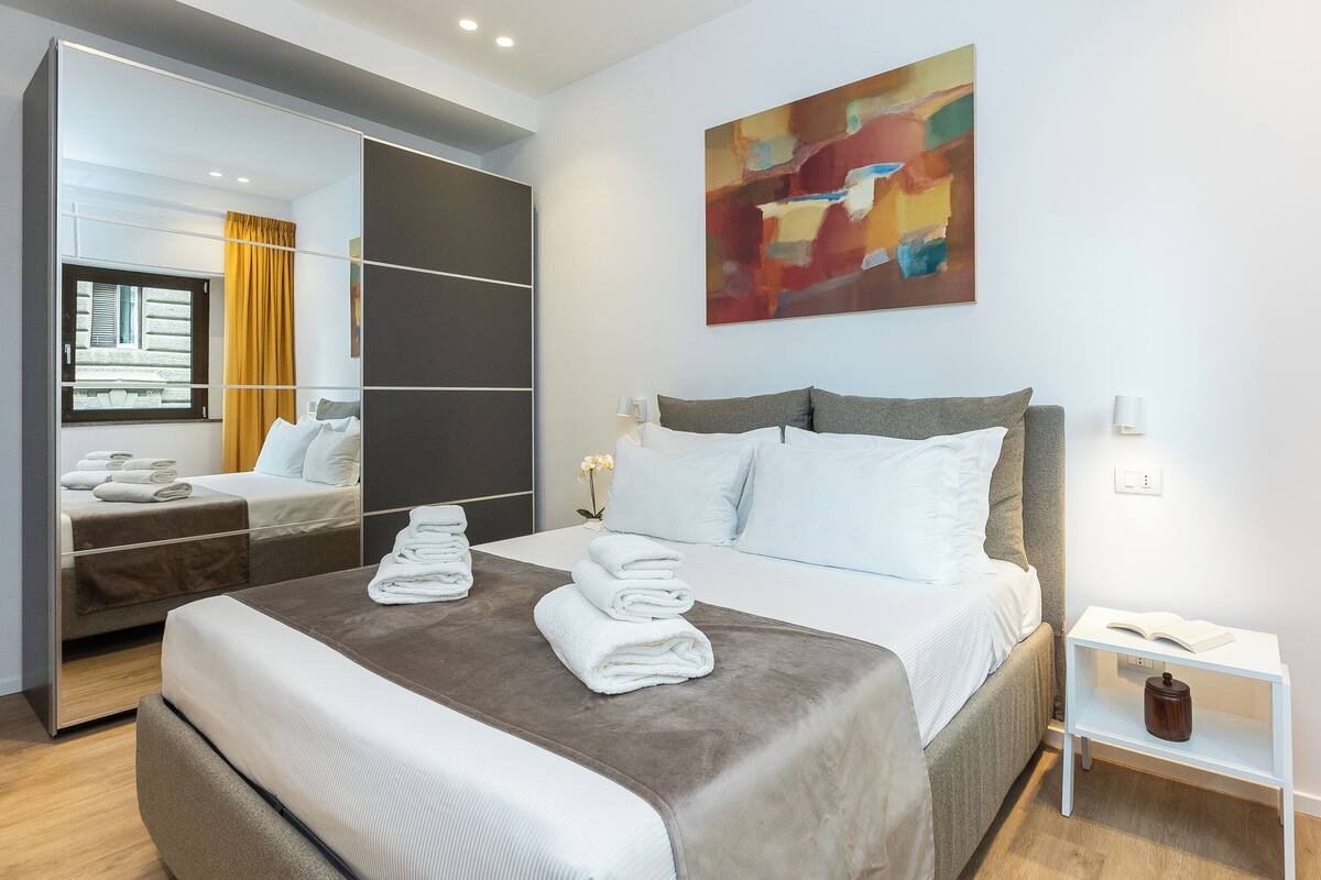 Ein modernes Hotelzimmer mit einem großen Bett, einem Nachttisch mit Lampe, einem Spiegel und einem farbenfrohen abstrakten Gemälde an der Wand.