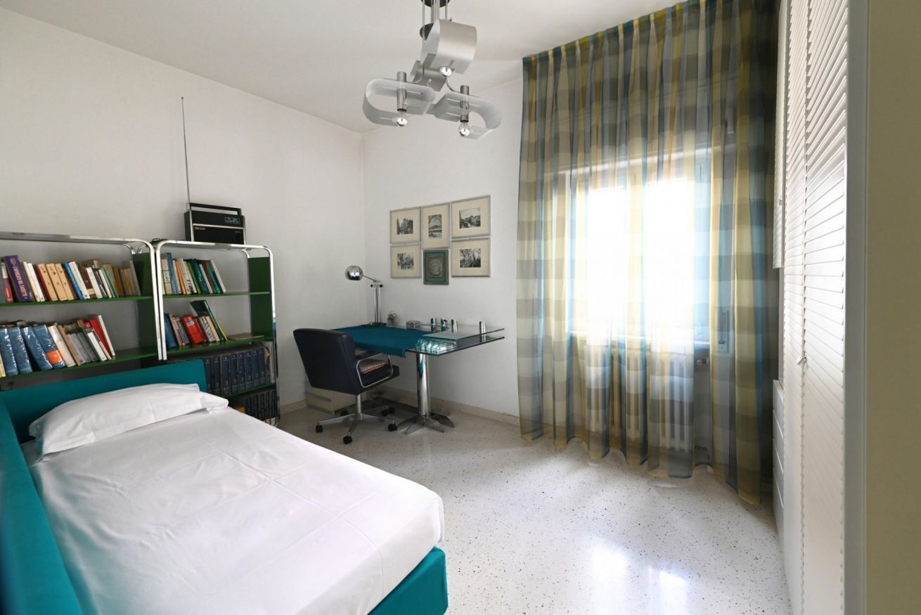A room with a bed, a desk, a chair, a bookshelf, and a window with curtains.