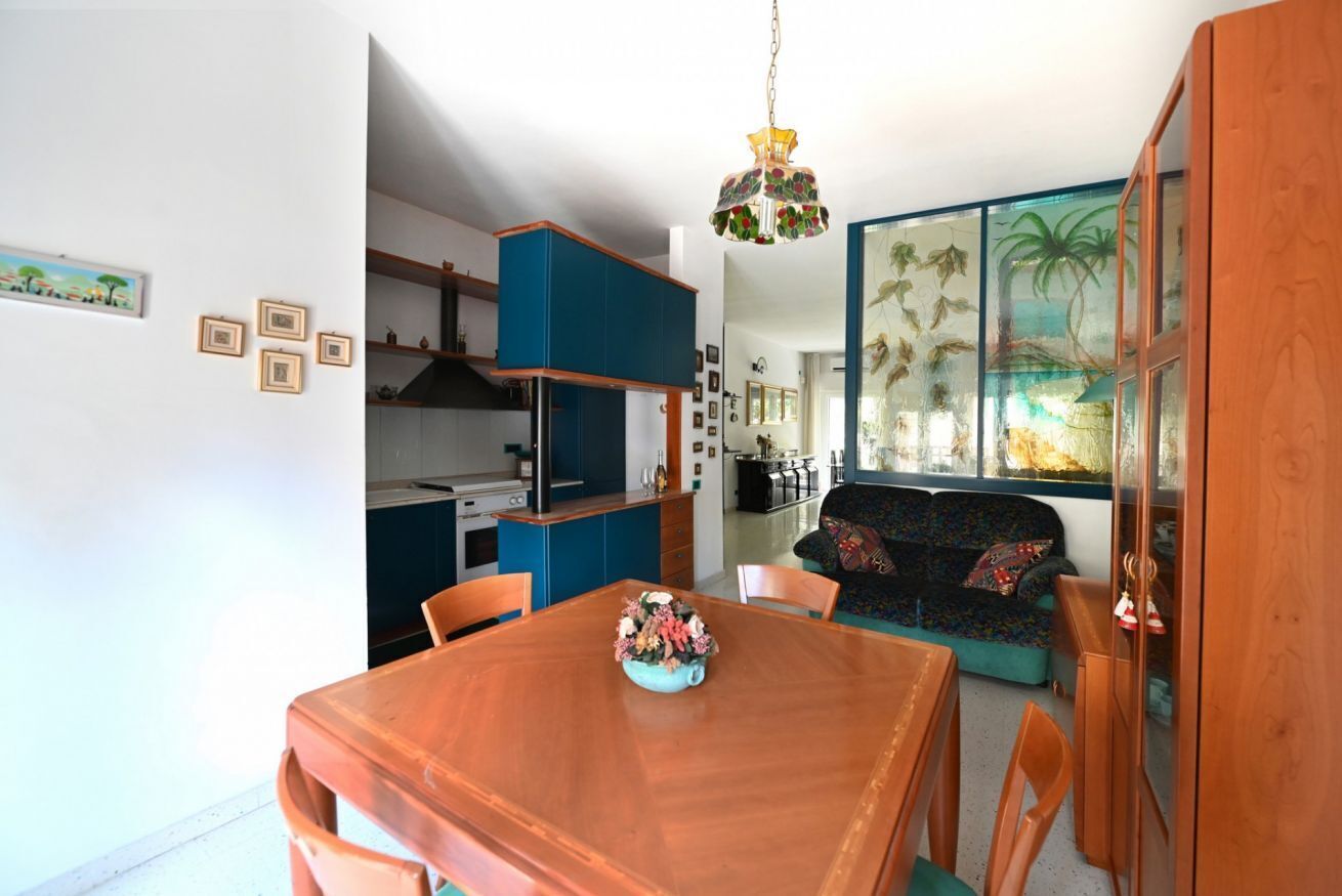 Un coin repas avec une table et des chaises en bois, une cuisine avec des meubles bleus et un salon avec un canapé et une grande fenêtre ornée d’une fresque aux motifs tropicaux.