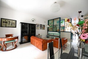Una sala de estar con un sofá, una mesa de comedor y sillas. Hay una araña, obras de arte en las paredes y un florero con flores.