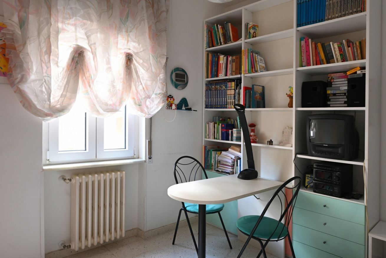 Una stanza con un termosifone, un tavolo con sedie, una libreria piena di libri, una televisione e una finestra con tende leggere.