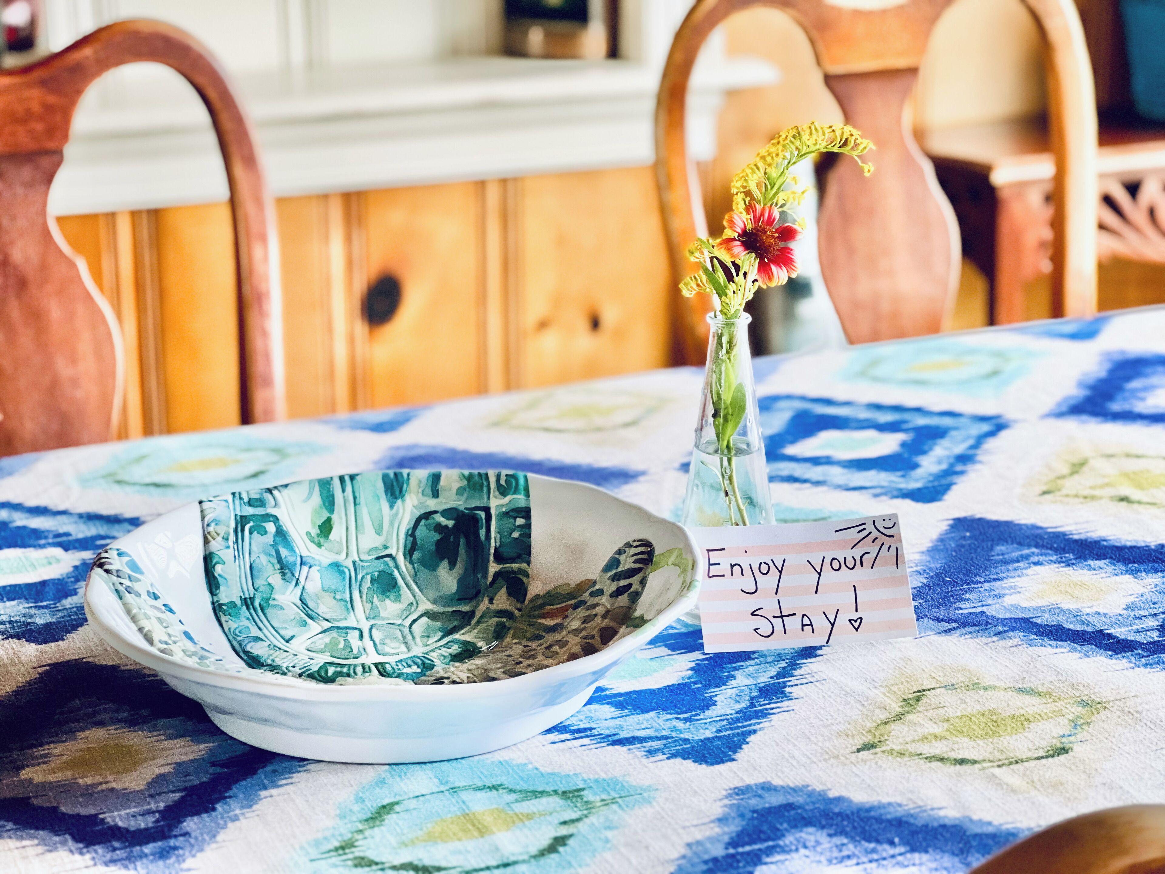 A dining table with a blue and white patterned tablecloth, a glass vase with a flower, and a sign that reads "Enjoy your stay!"