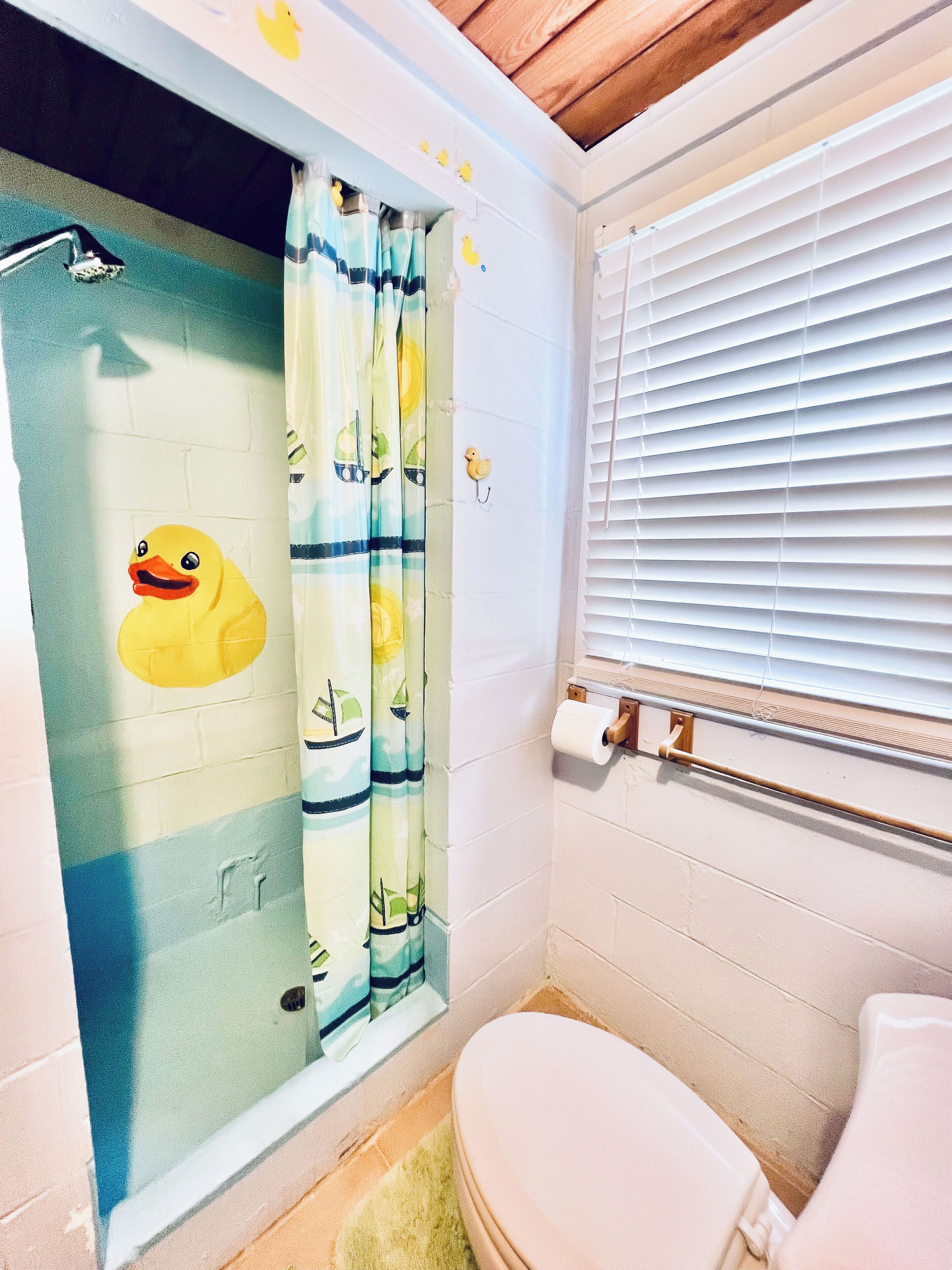 A bathroom with a shower curtain featuring a duck design, a toilet, and a window with blinds.