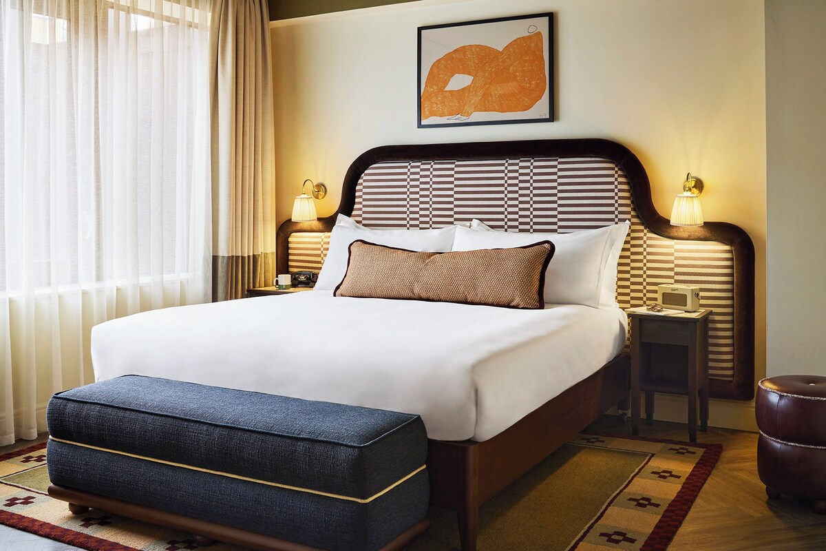 A hotel room with a large bed, a headboard with a checkered pattern, two bedside lamps, a framed artwork above the bed, a small table with a cup and a remote control, and a dark blue ottoman.