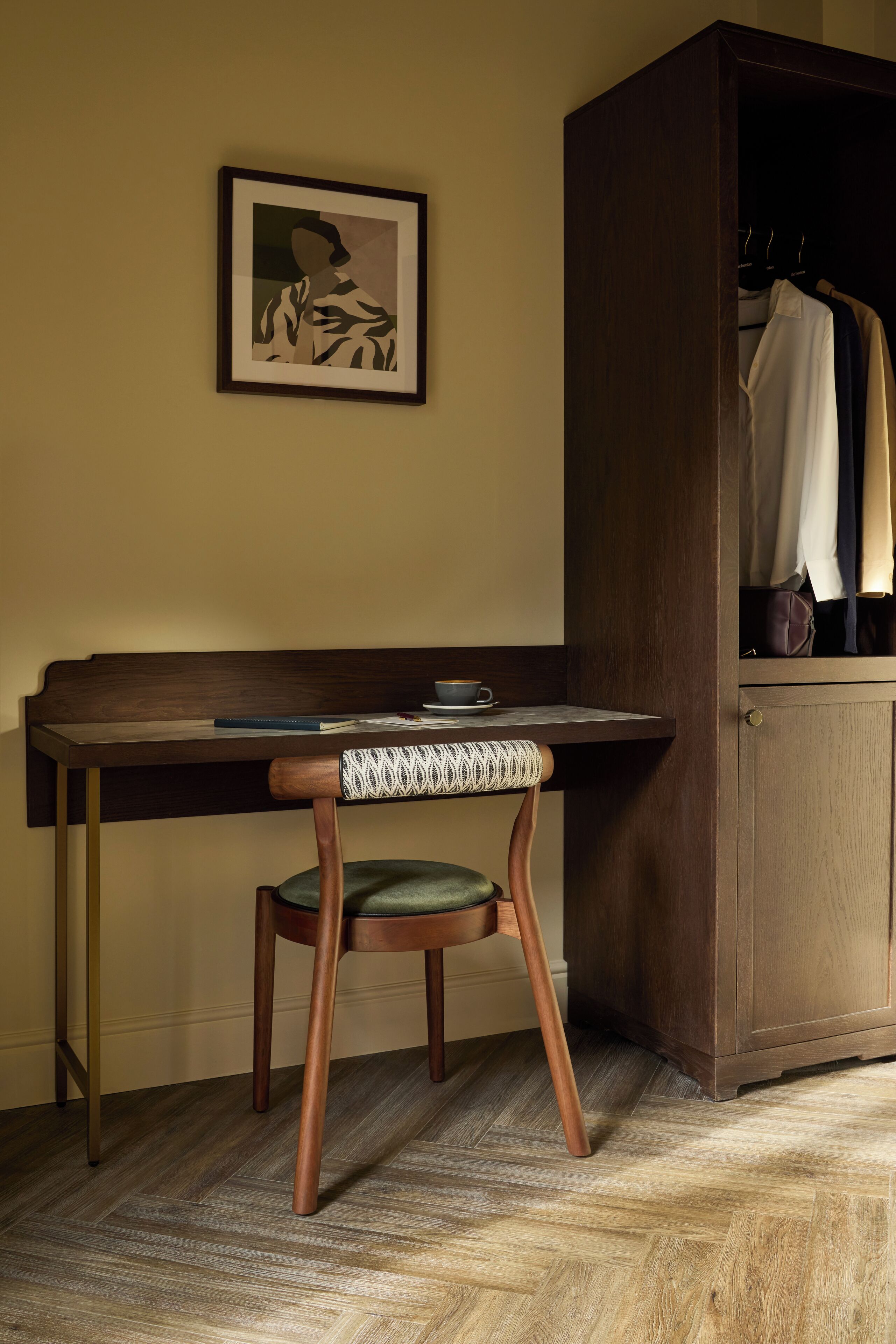 A wooden desk with a chair, a framed picture on the wall, and a wardrobe with hanging clothes.