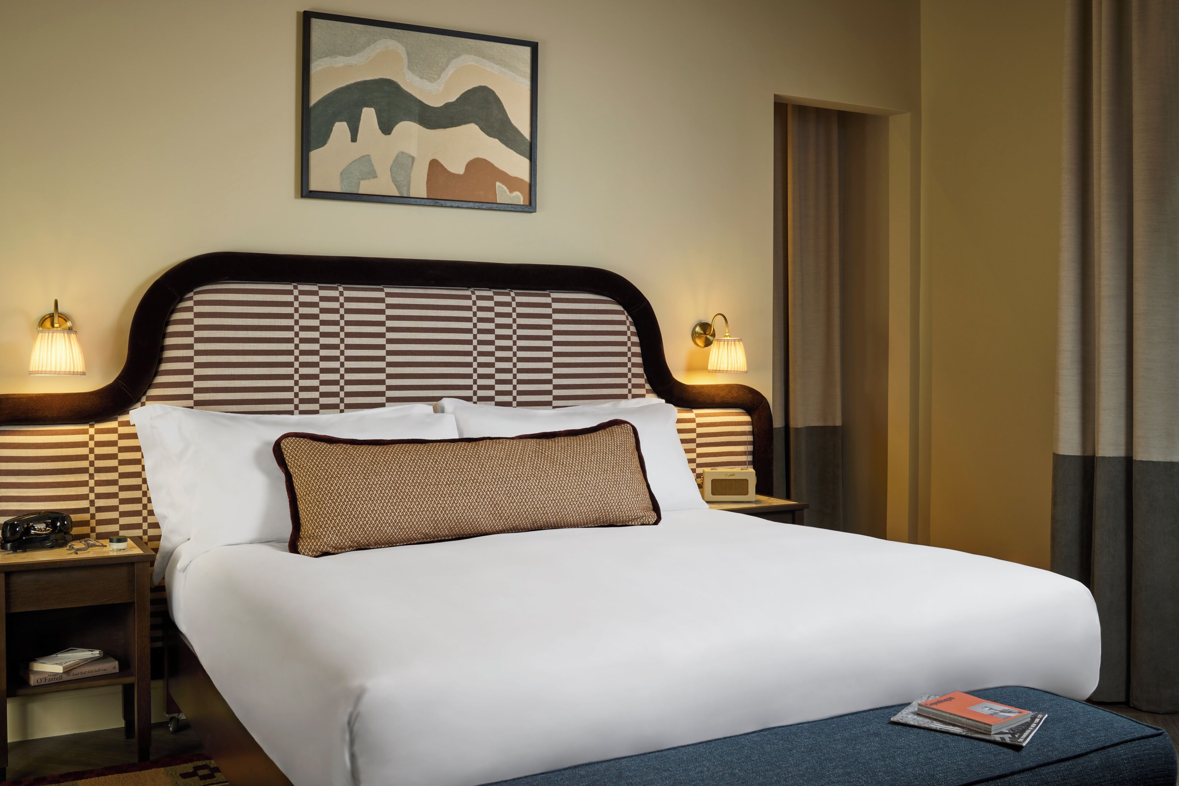 A neatly made bed with a checkered headboard, two bedside lamps, a framed artwork above the bed, and a magazine on the bed.