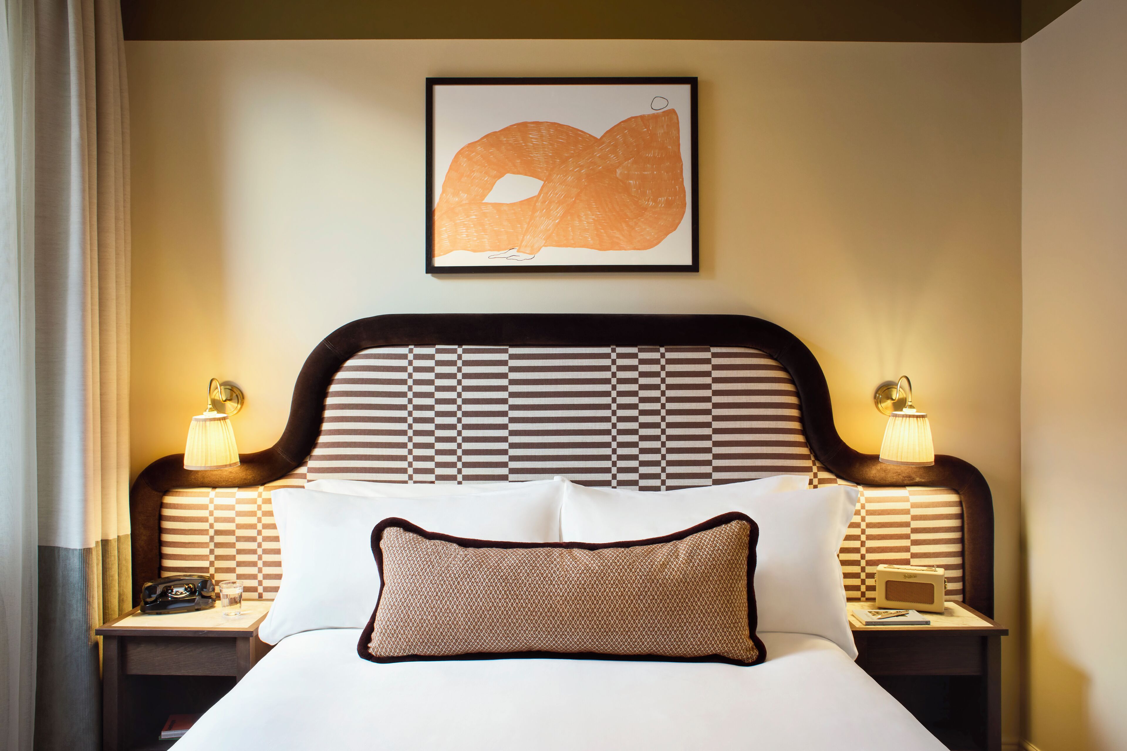 Une chambre d’hôtel avec un grand lit, deux tables de chevet, une lampe fixée au mur et une œuvre d’art encadrée au-dessus du lit.