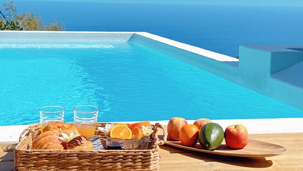 A poolside breakfast setup with fruits, pastries, and drinks, overlooking a clear blue sea.