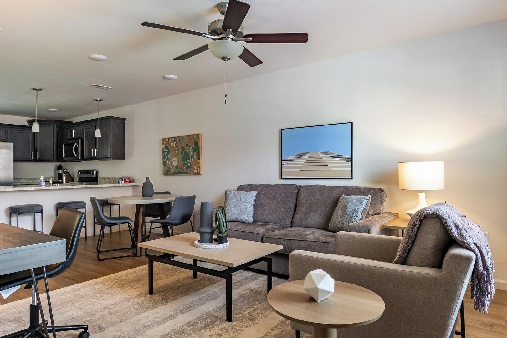 Ein modernes Wohnzimmer mit einer Couch, einem Couchtisch und Sesseln. Es gibt eine Küchenzeile mit Schränken und Geräten. Ein Deckenventilator ist sichtbar, und Kunstwerke sind an den Wänden ausgestellt.