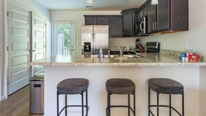 Una cocina moderna con armarios de madera oscura, electrodomésticos de acero inoxidable y una encimera de granito. Hay tres taburetes de bar y un bote de basura.