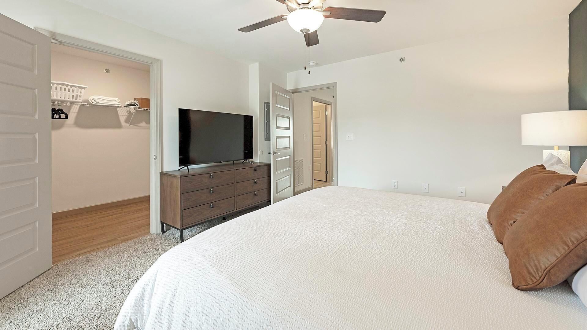 A bedroom with a bed, a TV on a dresser, a ceiling fan, and a closet with a laundry basket.