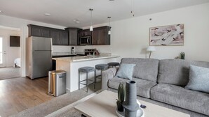 Una cocina moderna con armarios oscuros, un refrigerador de acero inoxidable y una encimera de mármol. En la sala de estar, hay un sofá gris con cojines estampados y un cactus sobre la mesa de centro.