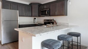 Cocina con armarios de madera oscura, refrigerador de acero inoxidable, microondas, estufa y encimera de granito. Hay tres taburetes de bar.