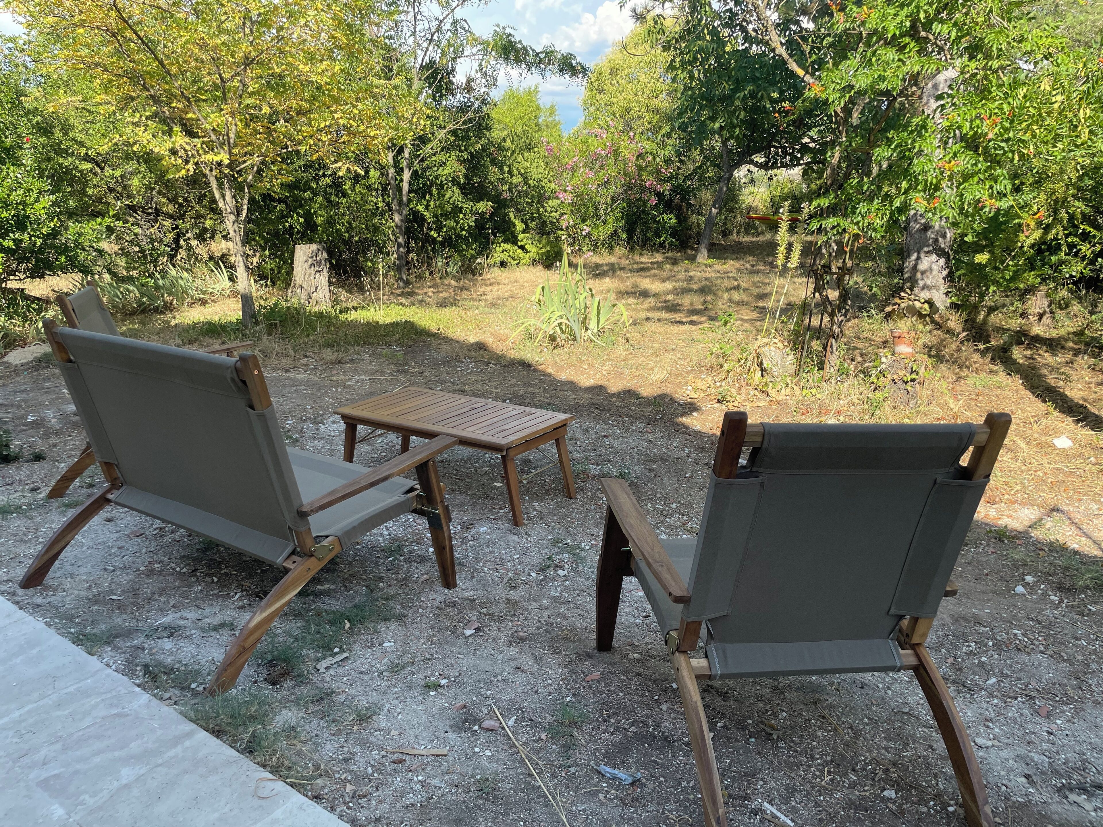 Two wooden-framed chairs and a small table in an outdoor setting with a gravel ground and greenery in the background.
