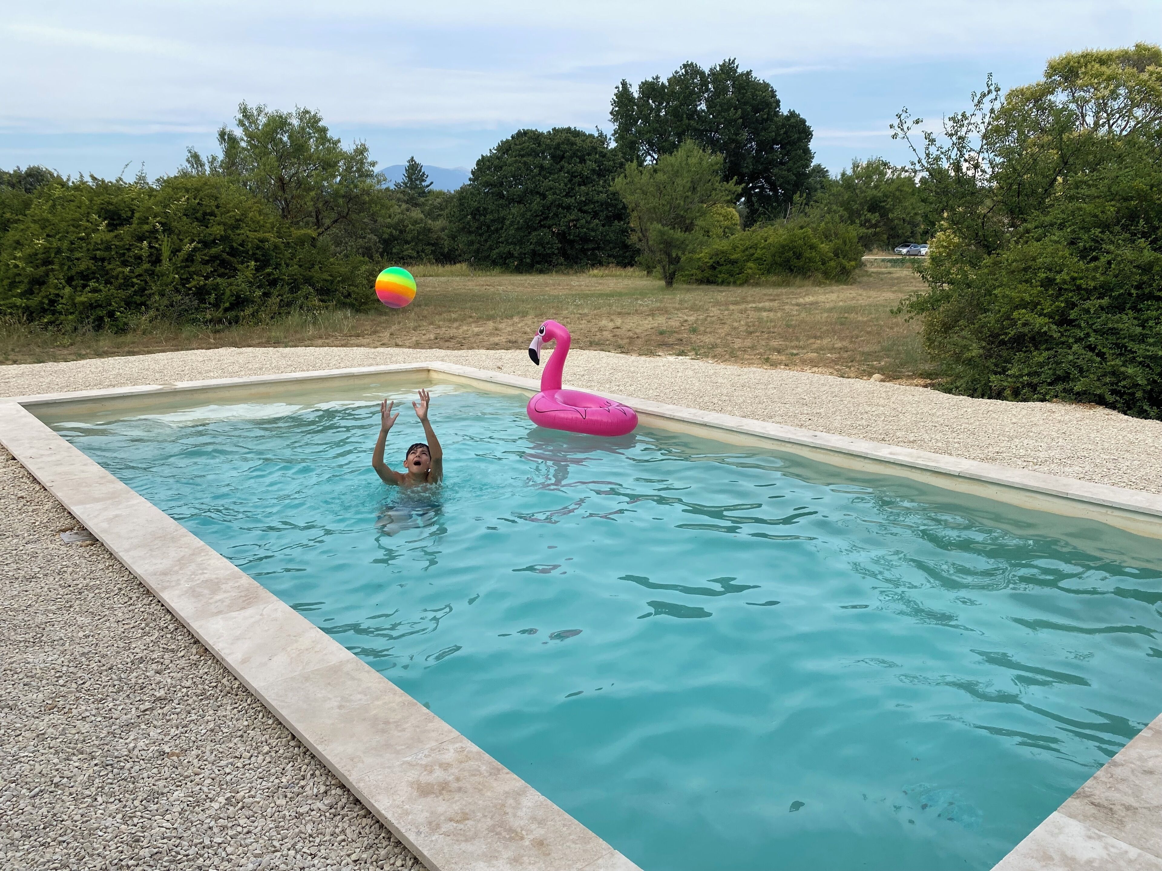 A swimming pool with a child playing with a ball, surrounded by pebbles and trees.