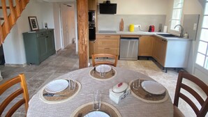 A dining area with a round table set for four, a kitchen in the background, and a wooden staircase leading to an upper level. - The farmhouse at the foot of the Ventoux (Carpentras)
