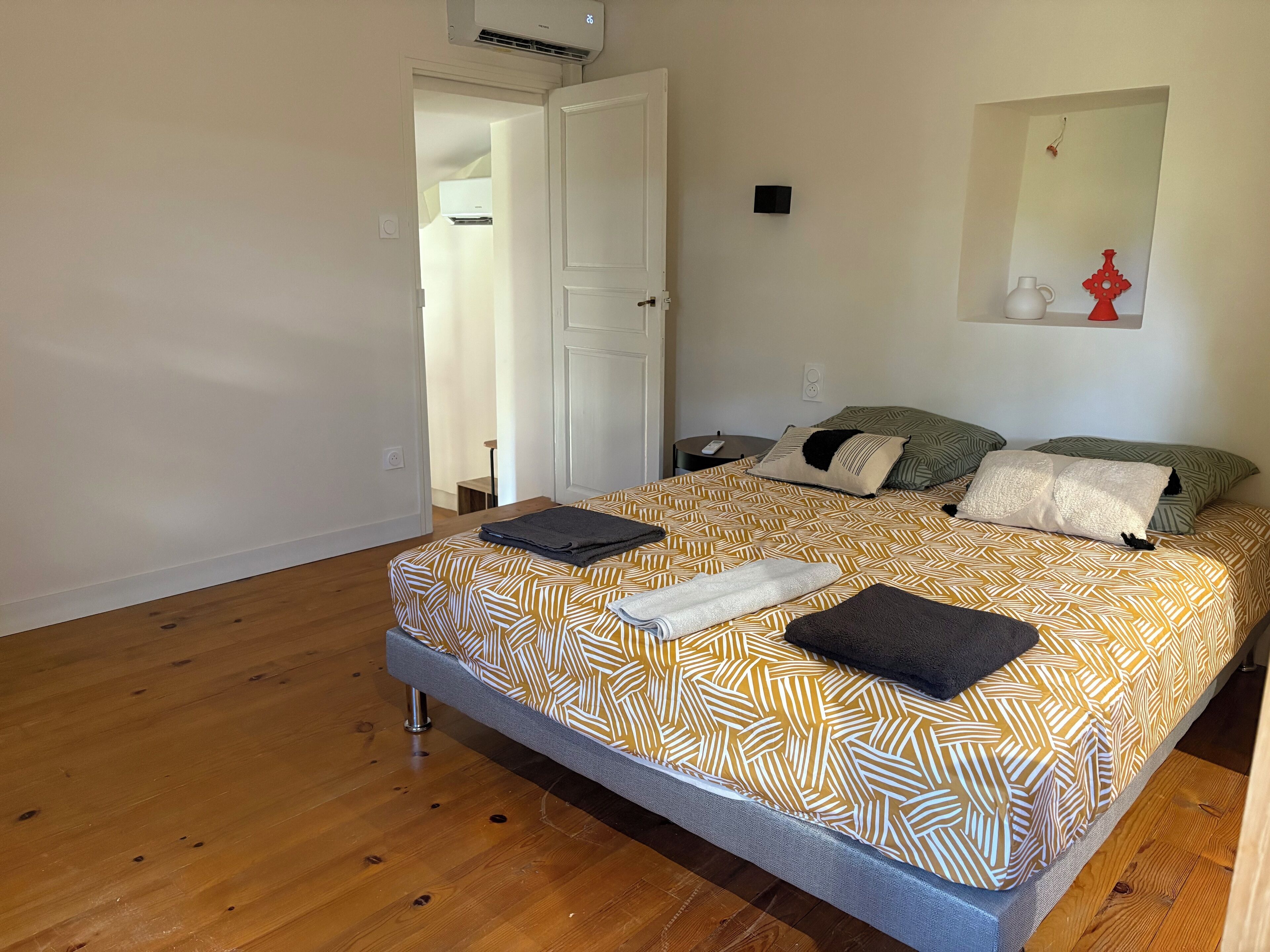 Un dormitorio con cama, mesita de noche, aire acondicionado instalado en la pared y una puerta que da a otra habitación.
