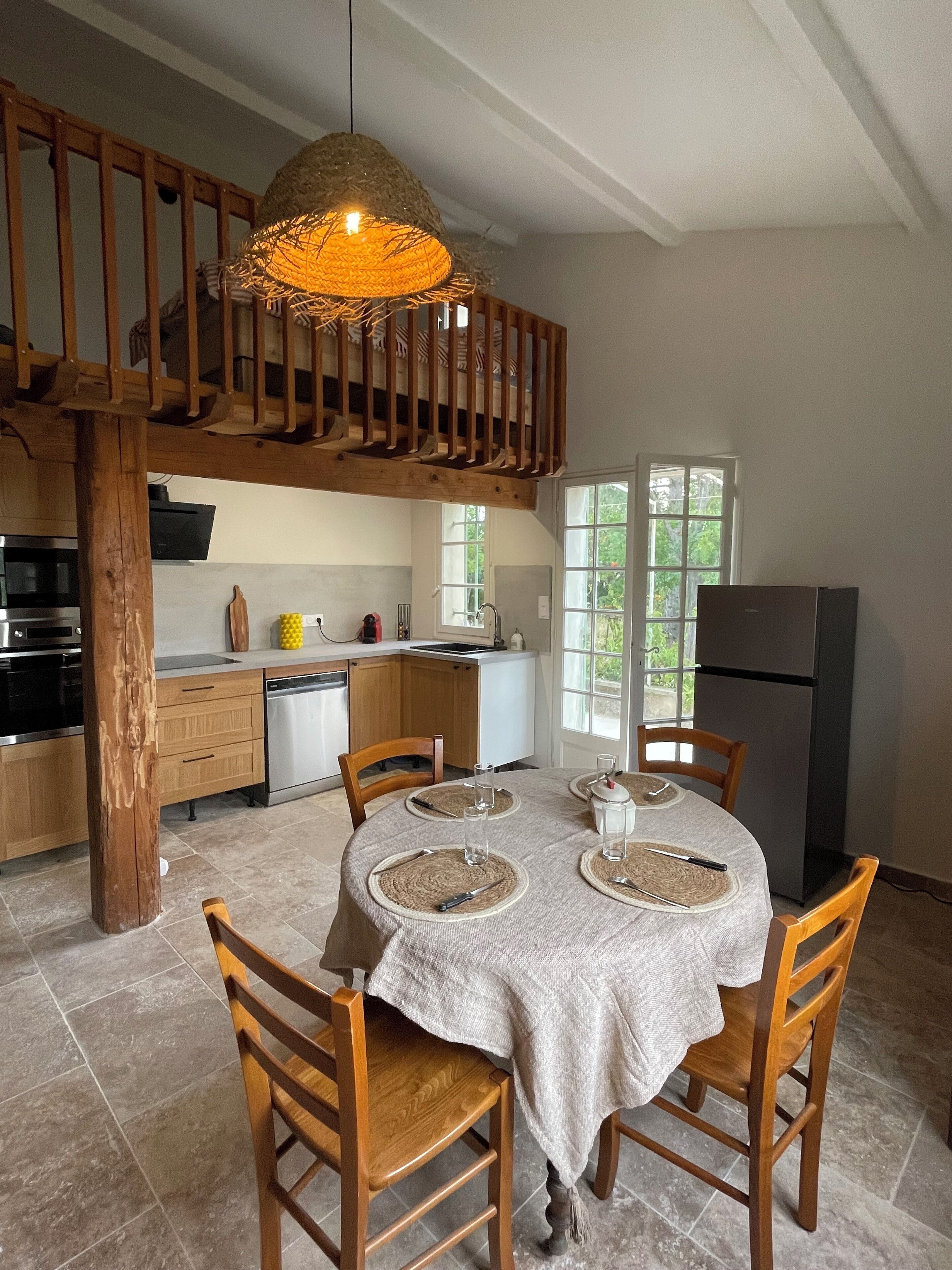 A dining area with a round table, wooden chairs, a hanging light fixture, and a kitchen in the background.