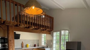 A dining area with a round table, wooden chairs, a hanging light fixture, and a kitchen in the background. - The farmhouse at the foot of the Ventoux (Carpentras)
