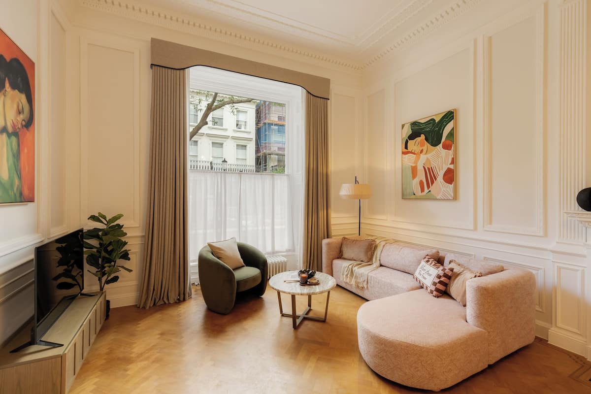 Sala de estar com sofá, poltrona e mesa de centro. Há uma janela com cortinas, uma planta e uma obra de arte na parede.