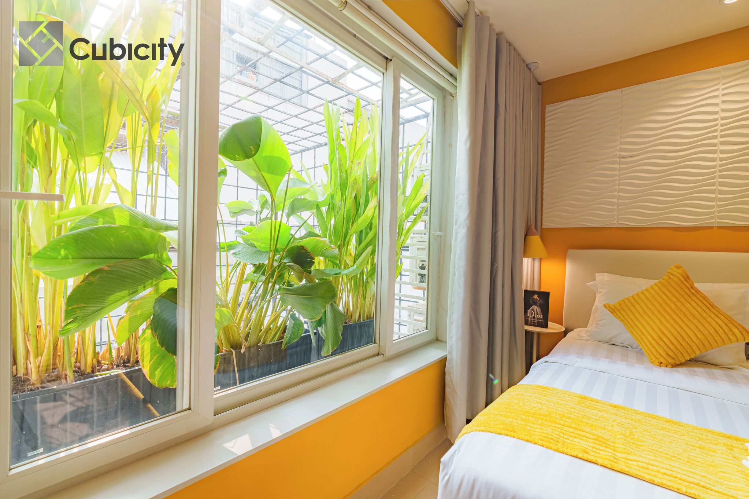 A hotel room with a large window, a bed with a yellow throw, and a nightstand with a lamp.