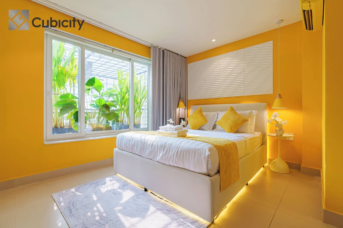 A modern bedroom with a large bed, a bedside table, a window with plants, and a view of greenery.