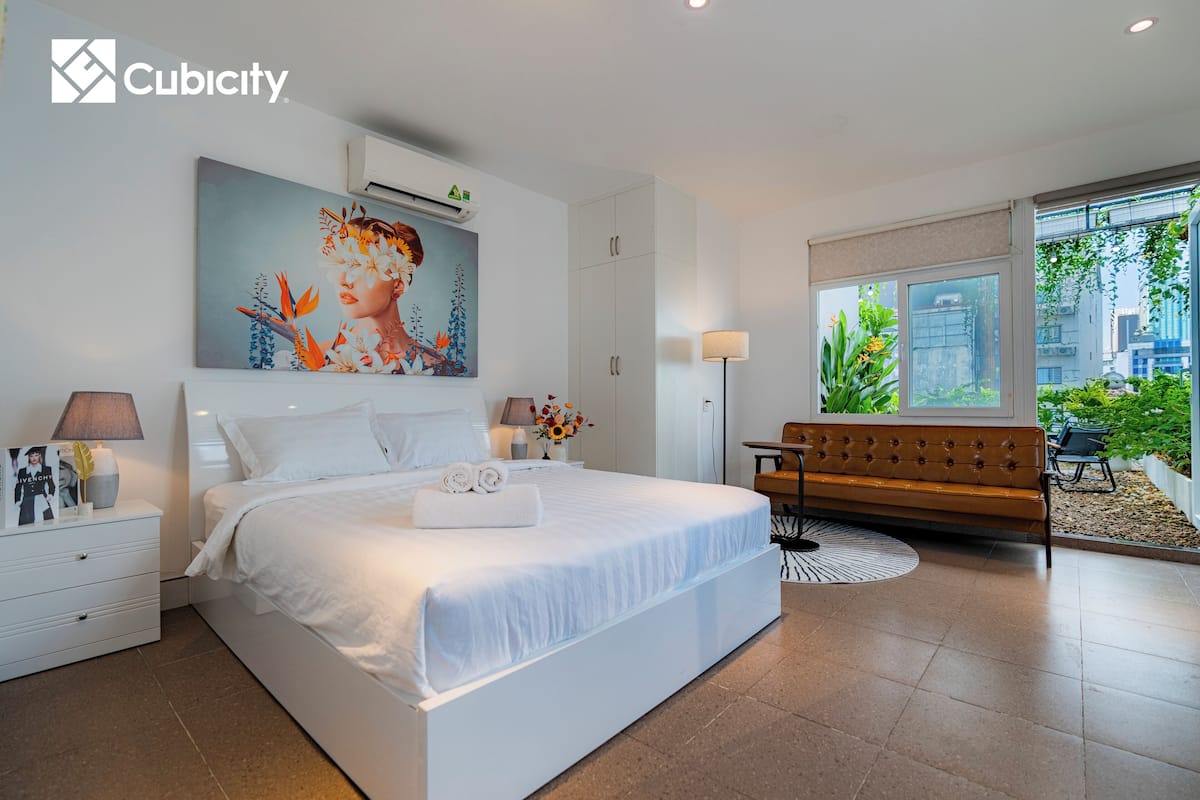 A modern hotel room with a large bed, a sofa, a desk, and a window with a view of greenery.