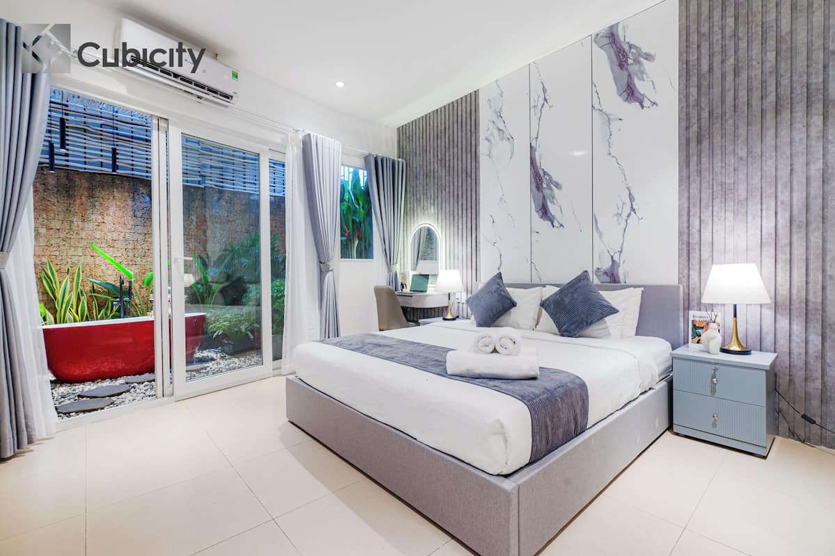A modern bedroom with a large bed, a nightstand, a red planter, and a view of greenery through the glass door.